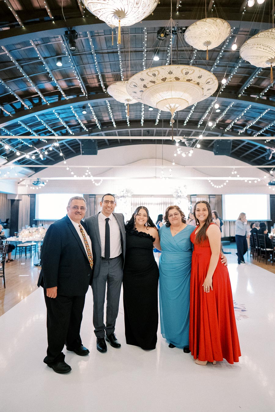 A group of five people dressed formally standing on a dance floor at a decorated indoor event venue with hanging lanterns and string lights.