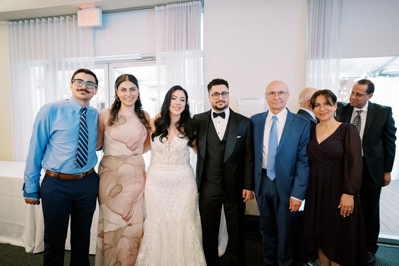 A group of people, including a bride in a white dress and a groom in a suit, standing together at an indoor wedding reception.