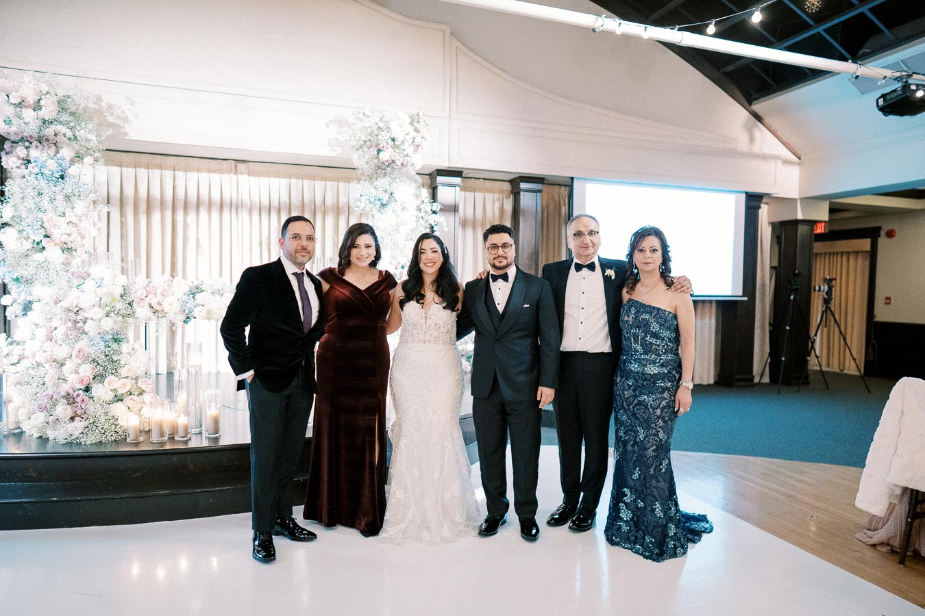 A group of six elegantly dressed people posing at a wedding reception, surrounded by floral decorations and candles, under ambient lighting in a venue.