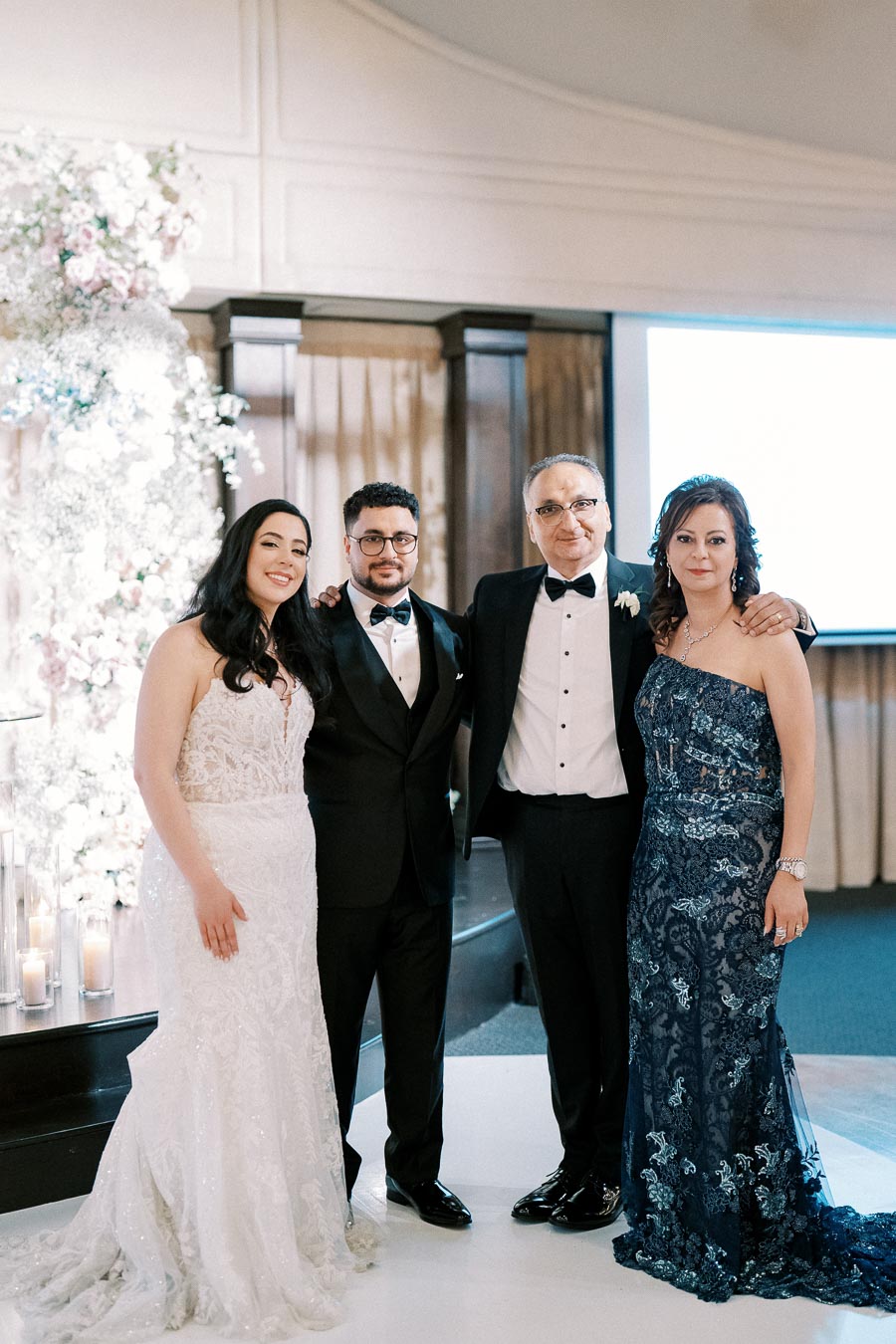 A bride in a white wedding gown stands next to a groom in a black tuxedo, accompanied by two elegantly dressed adults at a wedding reception, with a floral backdrop and romantic candles.