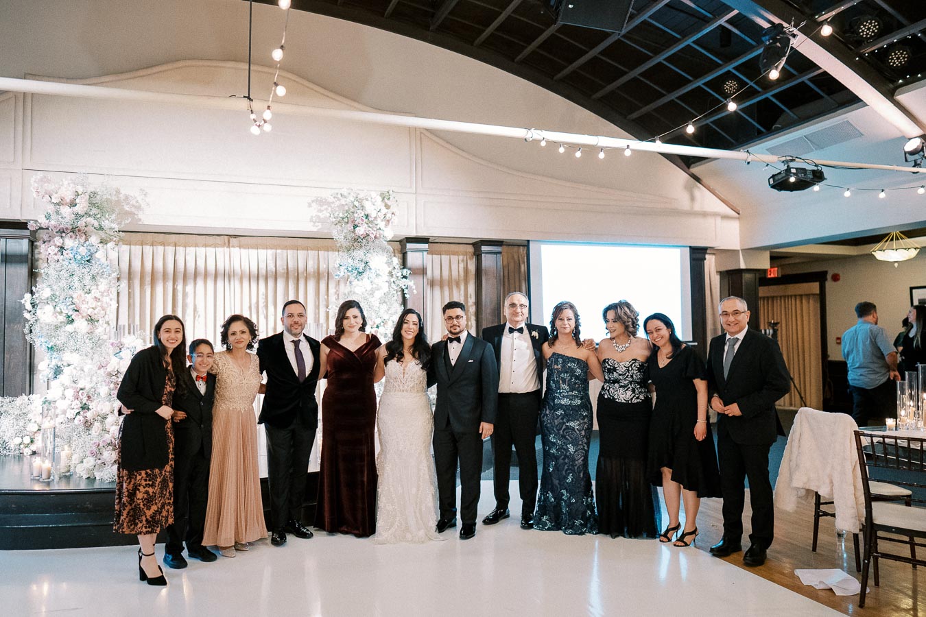 A group of people dressed in formal attire posing together at an elegantly decorated indoor event, featuring floral arrangements and soft lighting.