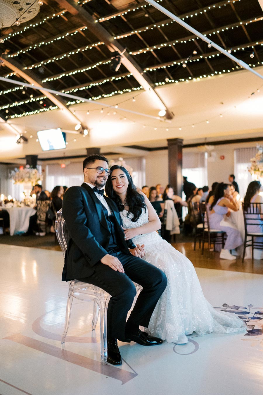 A newlywed couple in formal attire smiling as they sit together during their elegant wedding reception, surrounded by guests and warm decorative lighting.