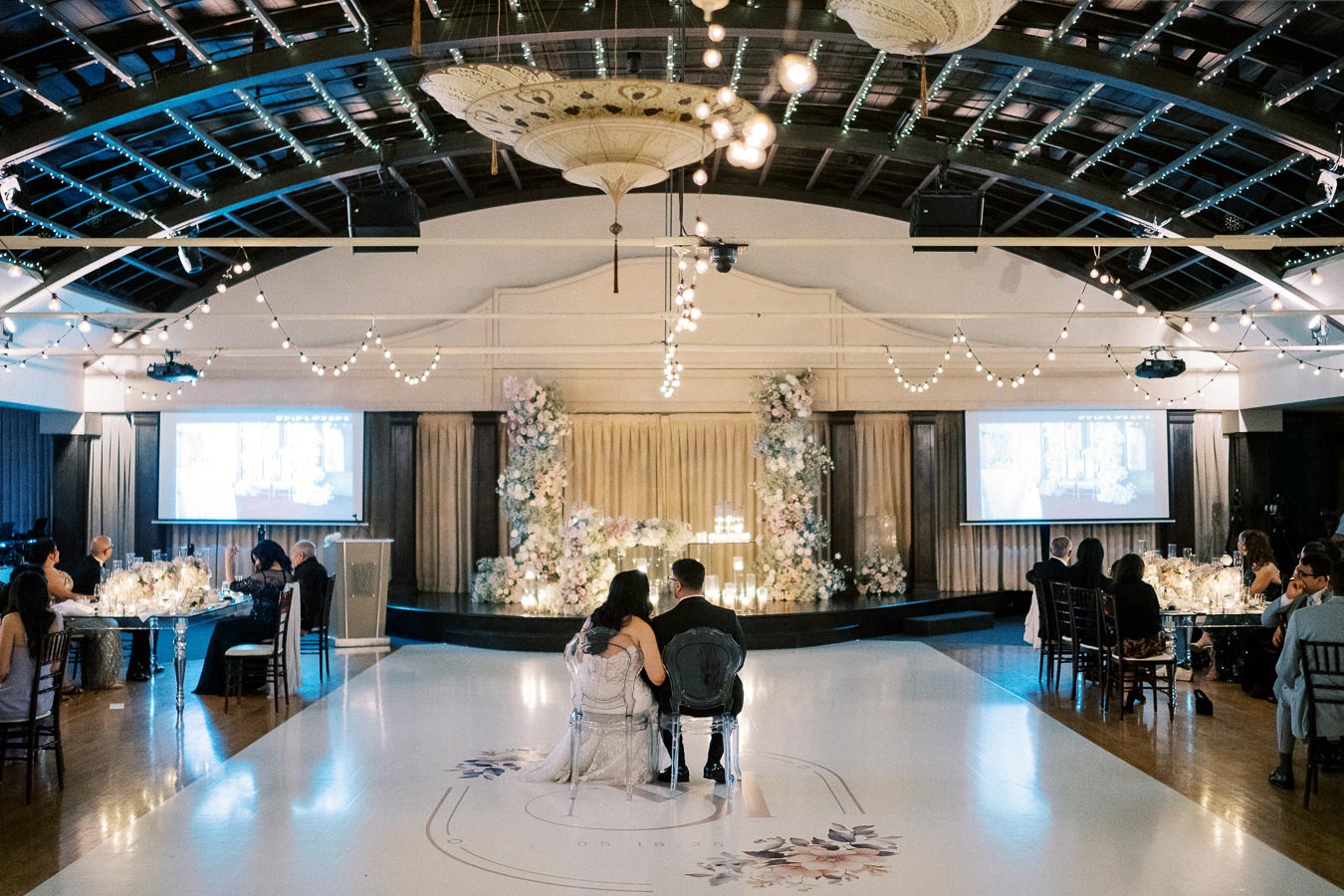 Wedding reception in an elegant venue with a couple sitting on a dance floor, surrounded by floral arrangements and string lights, while guests watch a slideshow on a large screen.