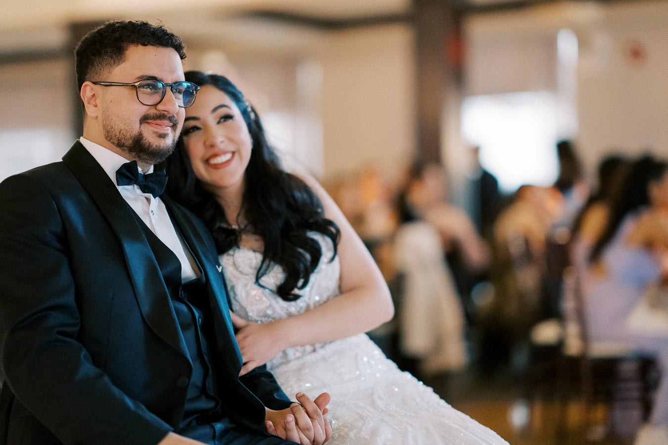 A happy couple sitting together at a wedding reception, with the bride in a white dress and the groom in a tuxedo, surrounded by guests.