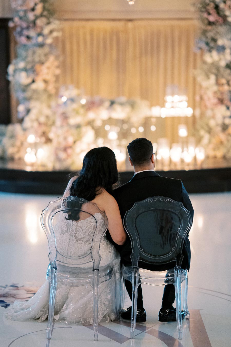 A bride and groom sit closely on transparent chairs, facing a beautifully decorated wedding altar adorned with flowers and candles, creating a romantic ambiance.