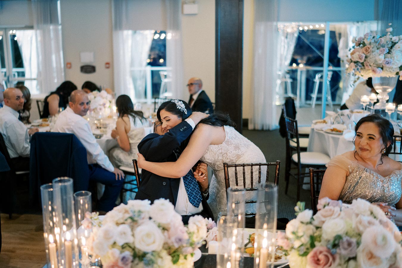 A joyful wedding reception scene with elegantly dressed guests seated around a beautifully adorned table. A woman in a white wedding dress lovingly embraces an older man in a suit, creating a heartwarming moment. The background is softly lit, showcasing floral arrangements and candlelight, enhancing the romantic atmosphere.