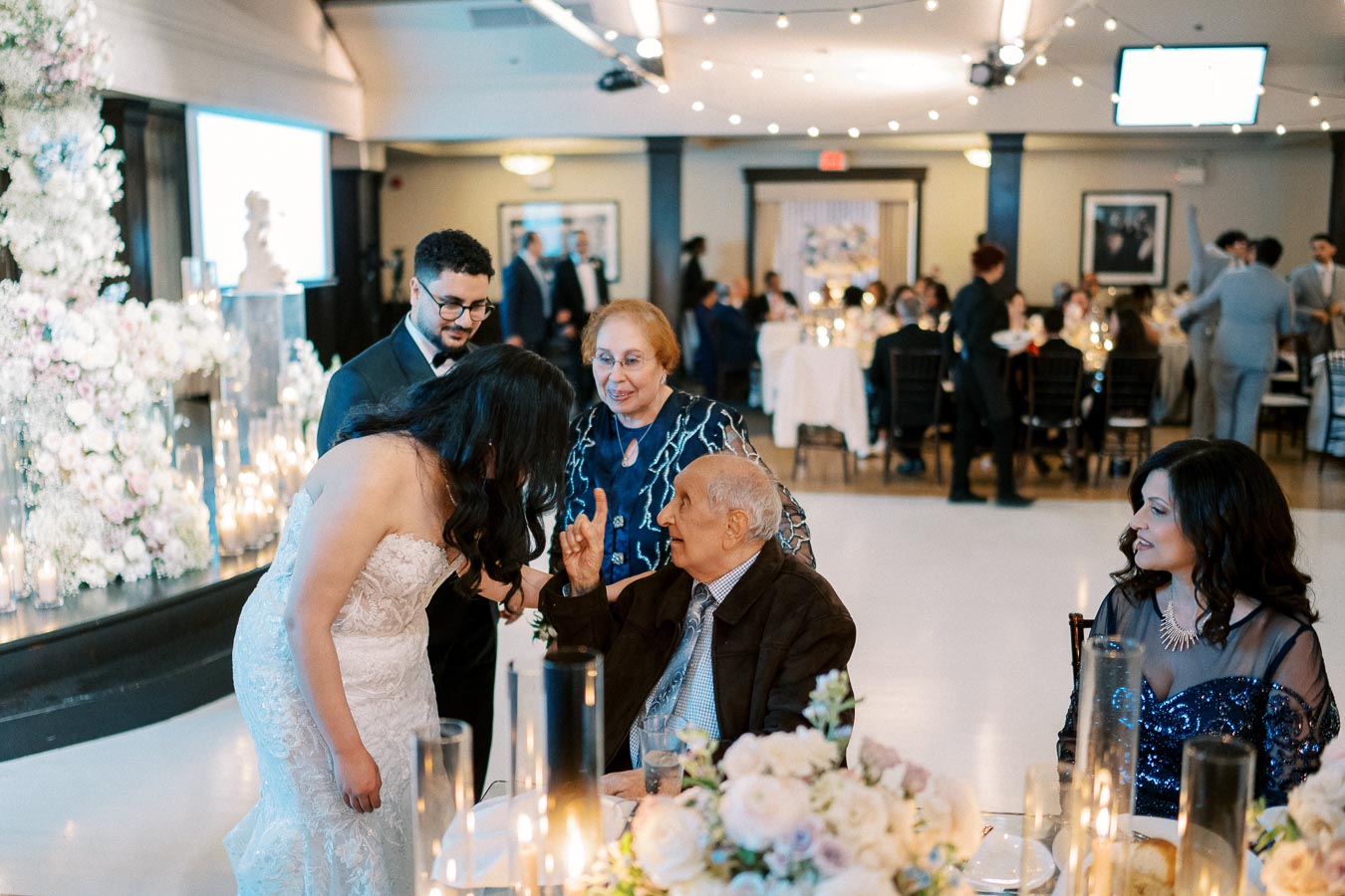 A bride in a white lace gown interacts with an elderly guest seated at a table during a wedding reception, surrounded by elegantly dressed attendees, floral arrangements, and candlelit decor.