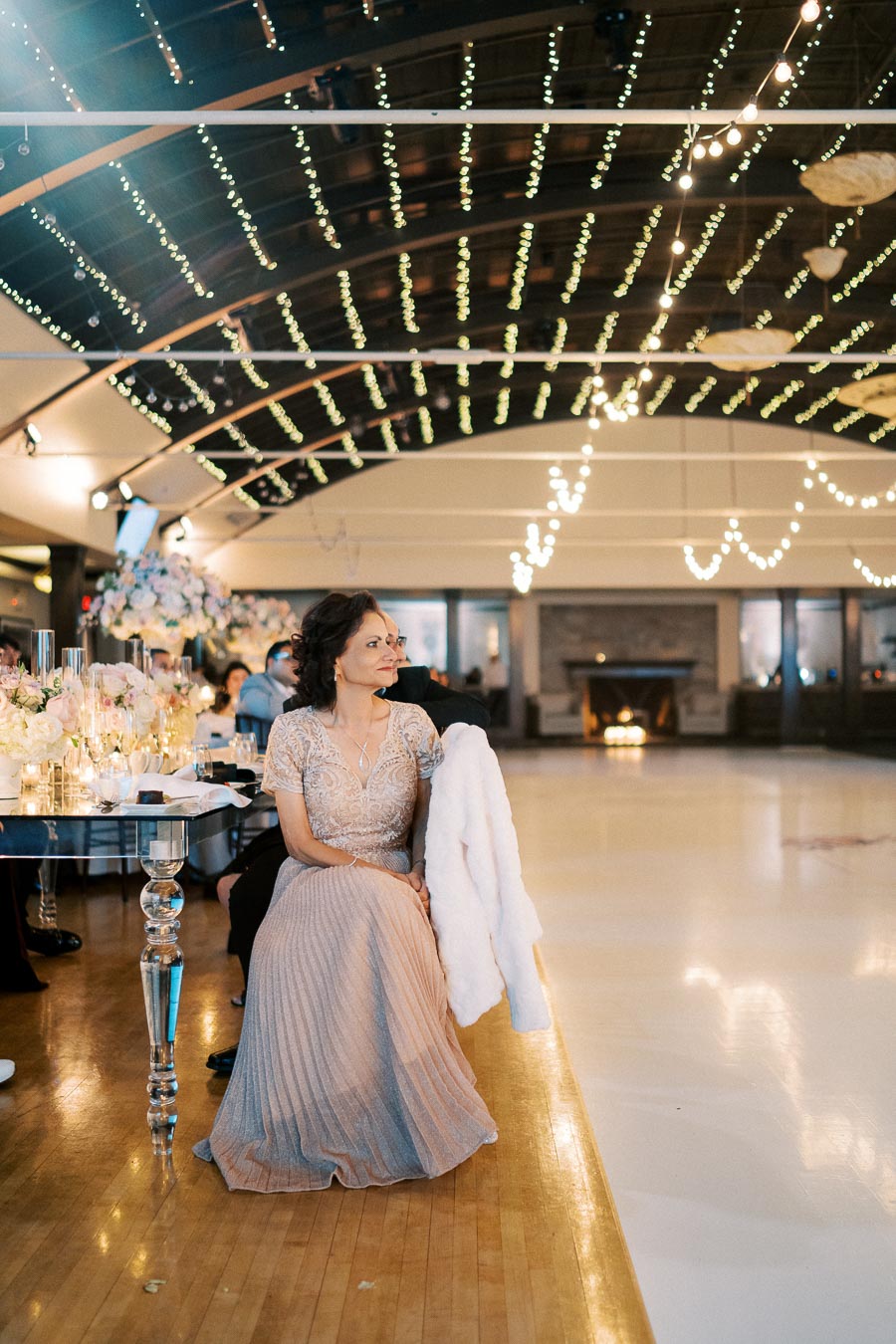 A woman in an elegant gown sits at a decorated table, surrounded by ambient string lights in a beautifully lit ballroom setting, creating a warm and celebratory atmosphere.