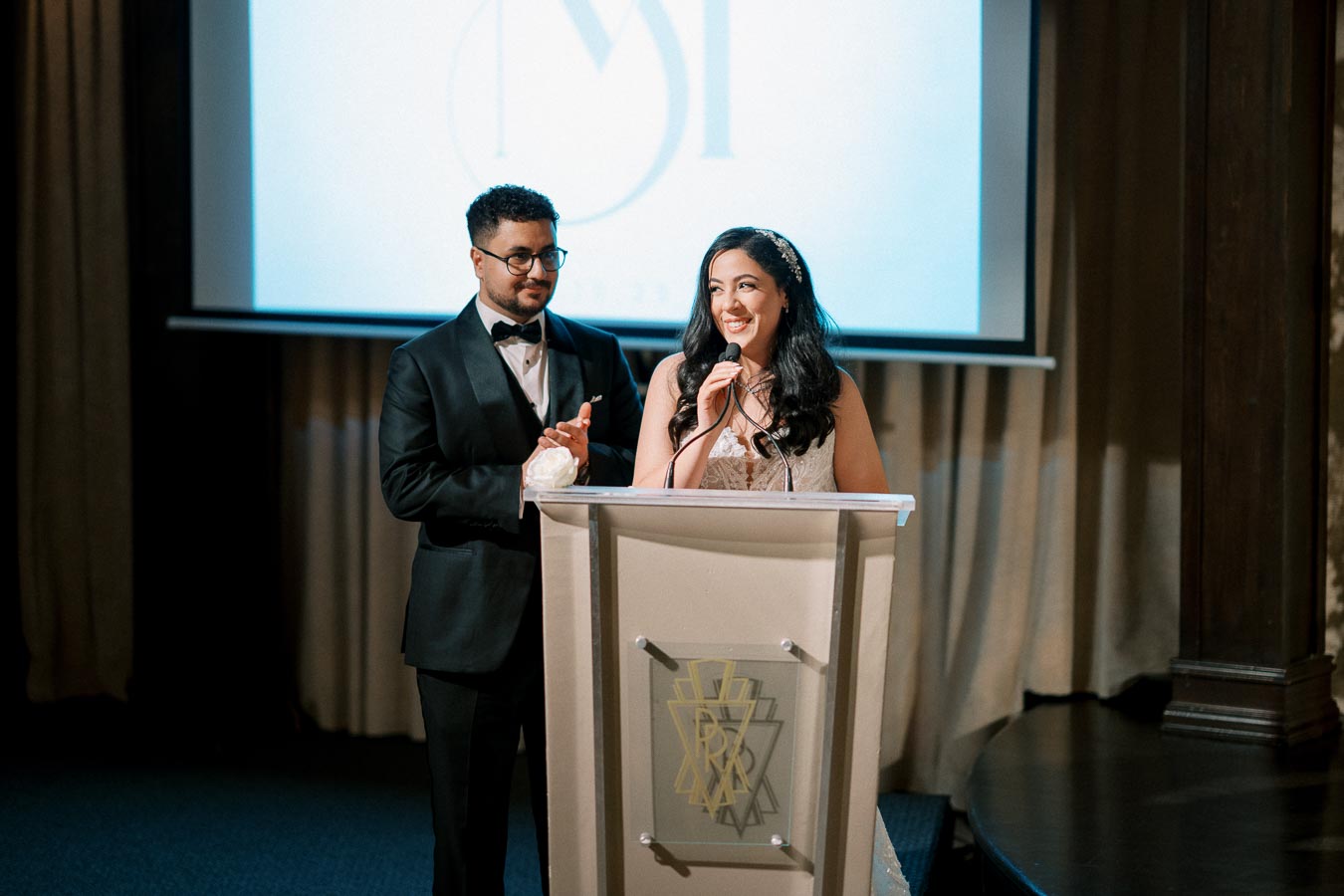 A bride and groom smiling and standing at a podium during a wedding reception speech, dressed in formal attire, with a projection screen in the background.