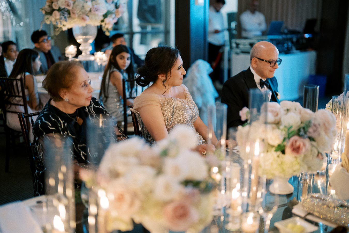 Elegant wedding reception with guests seated at a decorated table, featuring floral centerpieces and candlelight, creating a warm and inviting atmosphere.