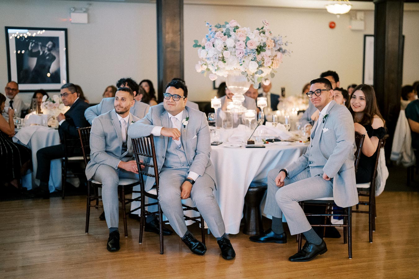 Guests in formal attire enjoy a beautifully decorated wedding reception, seated around a table adorned with elegant floral arrangements and candlelight.