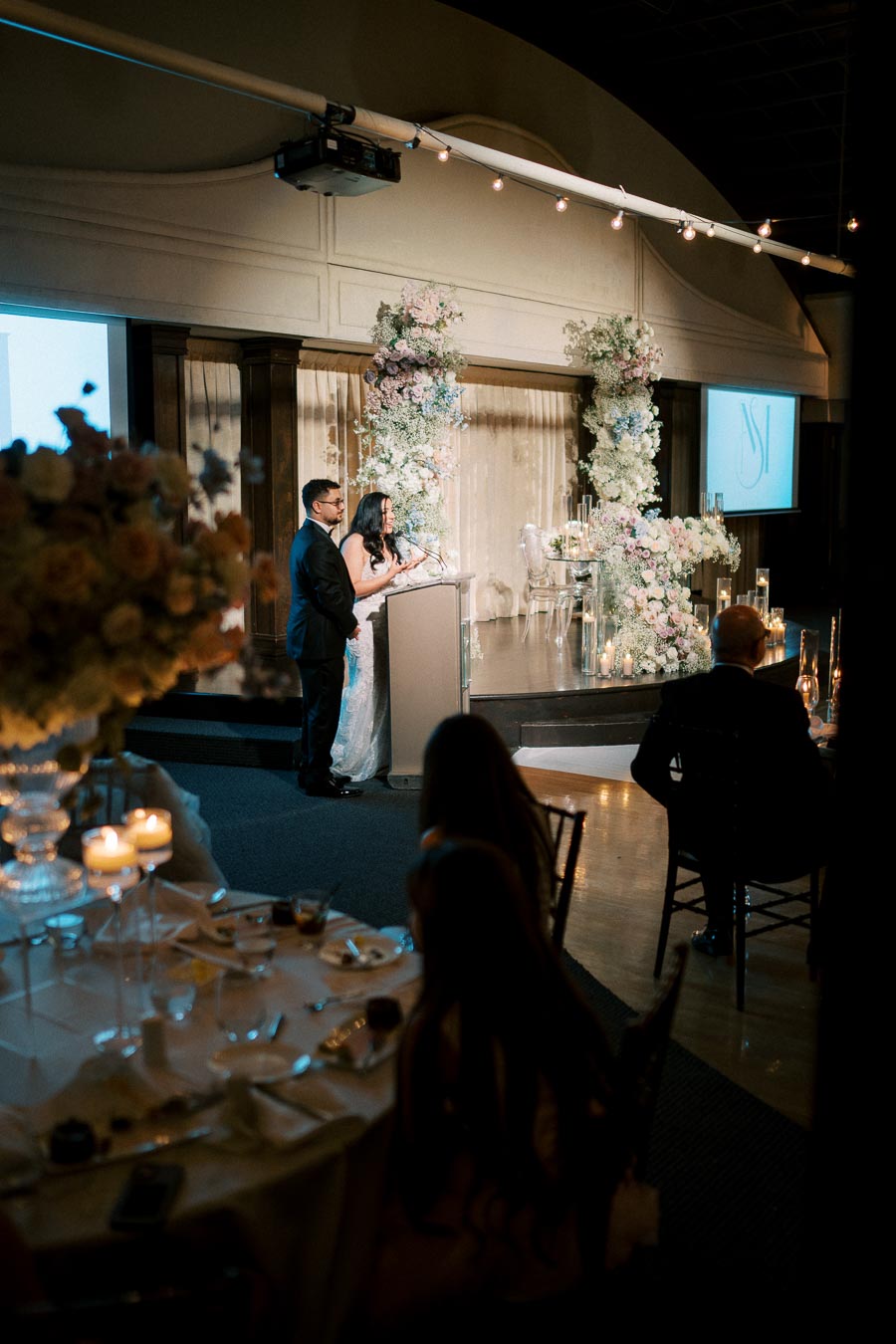 A couple delivering a speech at a beautifully decorated indoor wedding reception, surrounded by floral arrangements and candlelight ambiance.