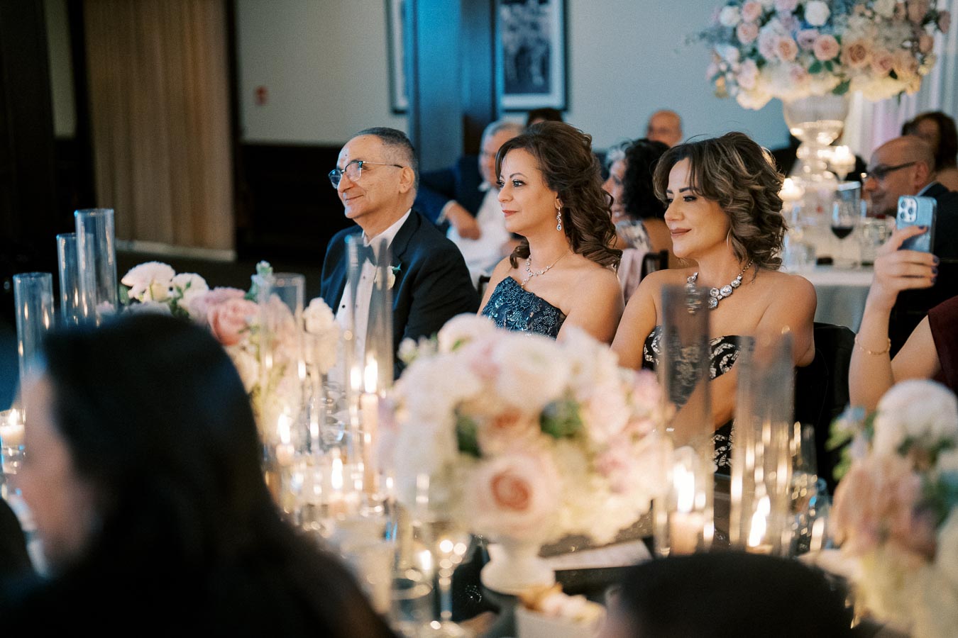 Elegant wedding reception with guests seated at a beautifully adorned table, featuring floral arrangements and candlelight, creating a warm and festive atmosphere.