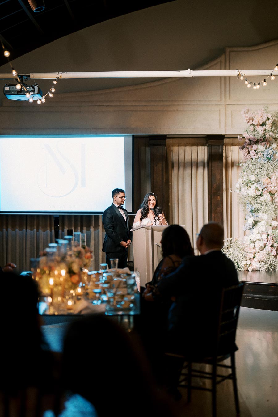 Wedding couple giving a speech at a reception, standing behind a podium with a floral backdrop and guests seated at candlelit tables in a warmly lit banquet hall.
