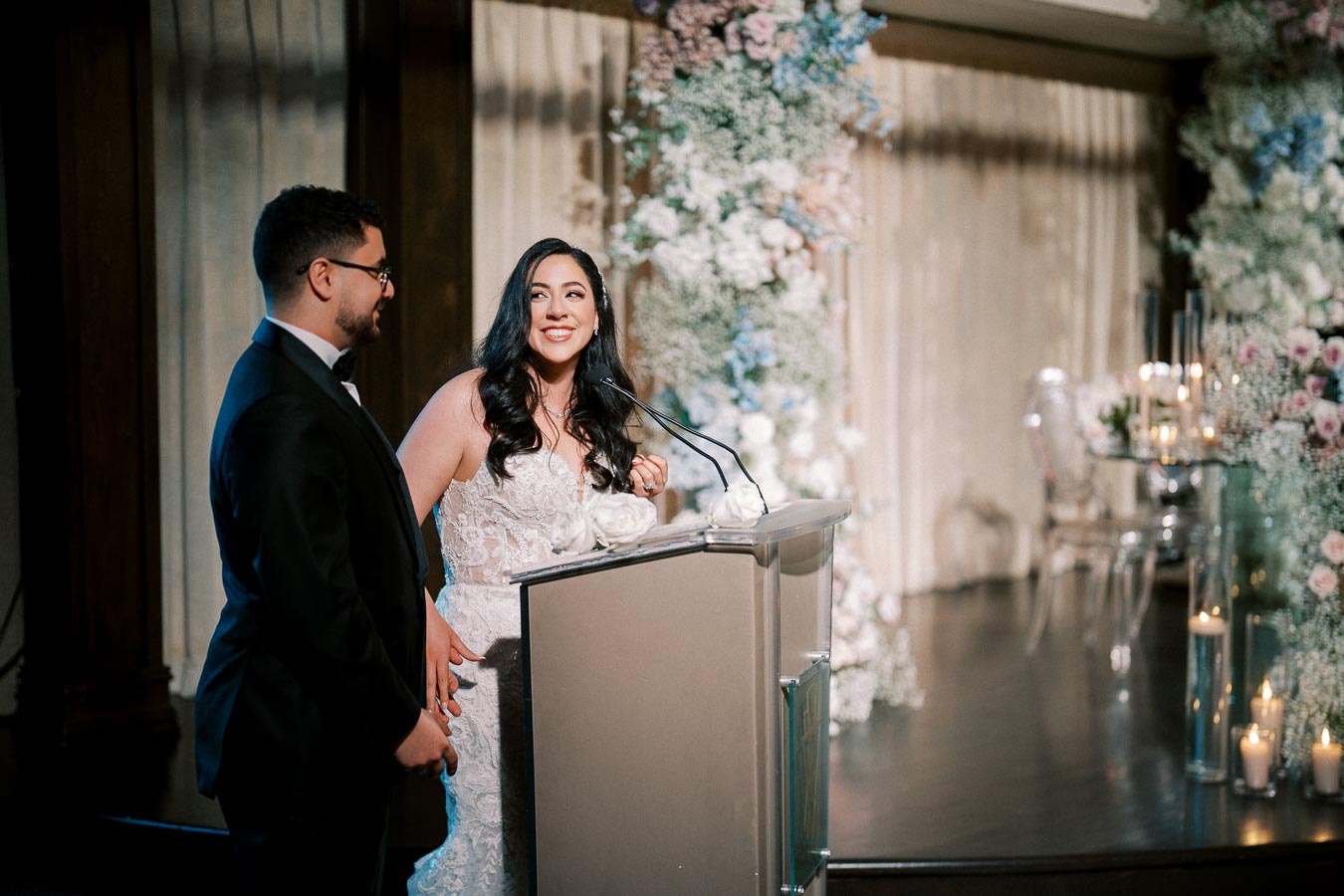 A bride and groom smiling while giving a speech at their wedding reception, standing behind a decorated podium with floral arrangements and lit candles in the background.