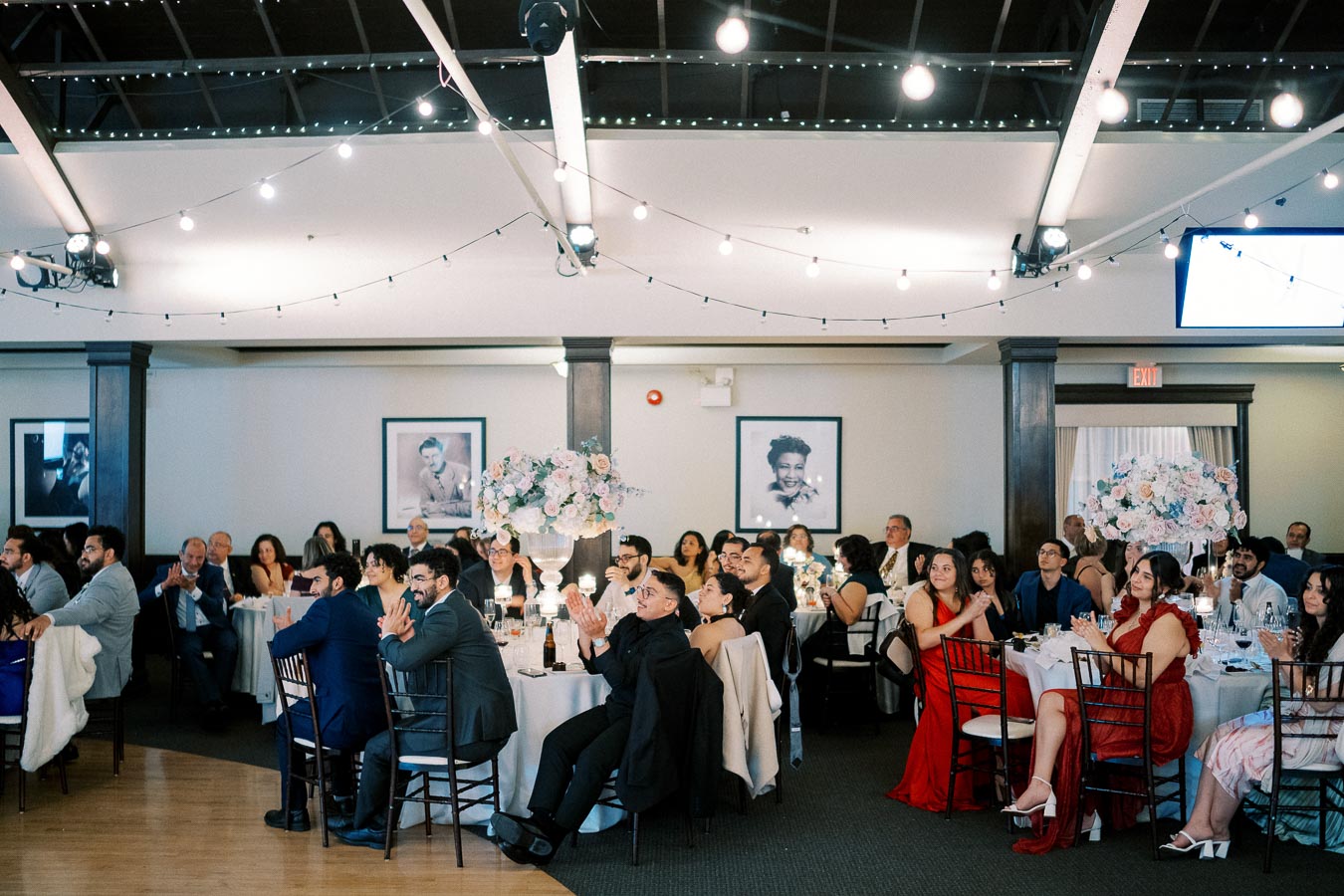 Elegant wedding reception with guests seated at round tables, adorned with floral centerpieces and string lights overhead in a cozy venue.