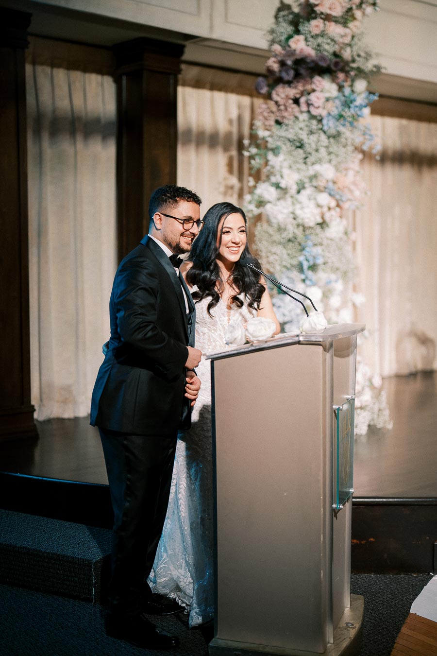A couple standing at a podium giving a speech during a wedding reception, with elegant floral arrangements in the background.