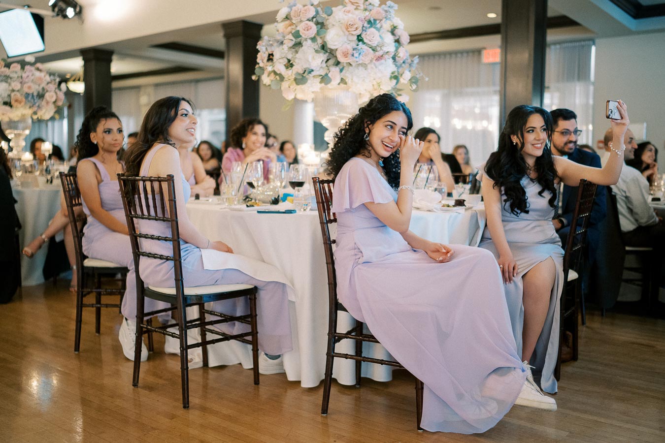 Wedding reception with smiling guests in lavender dresses seated at elegantly decorated table with floral centerpiece.