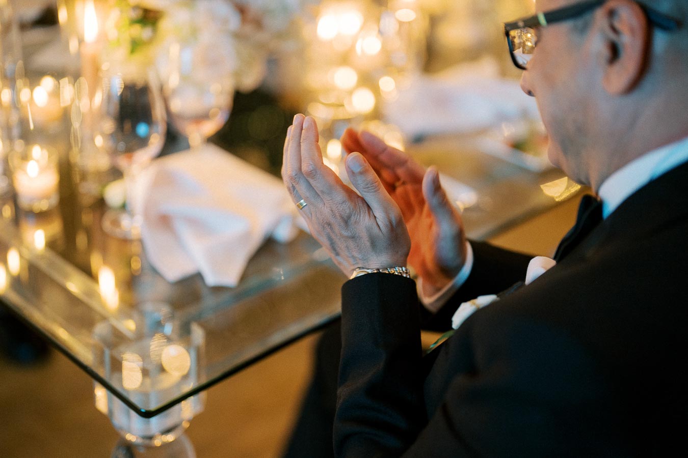 Man in a suit clapping at a formal event, seated at a candle-lit table with elegant glassware and floral decorations.