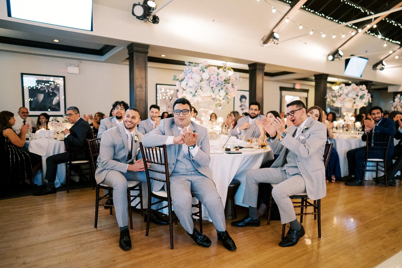Guests in elegant attire sit and applaud at a beautifully decorated wedding reception, with floral centerpieces and ambient lighting creating a festive atmosphere.