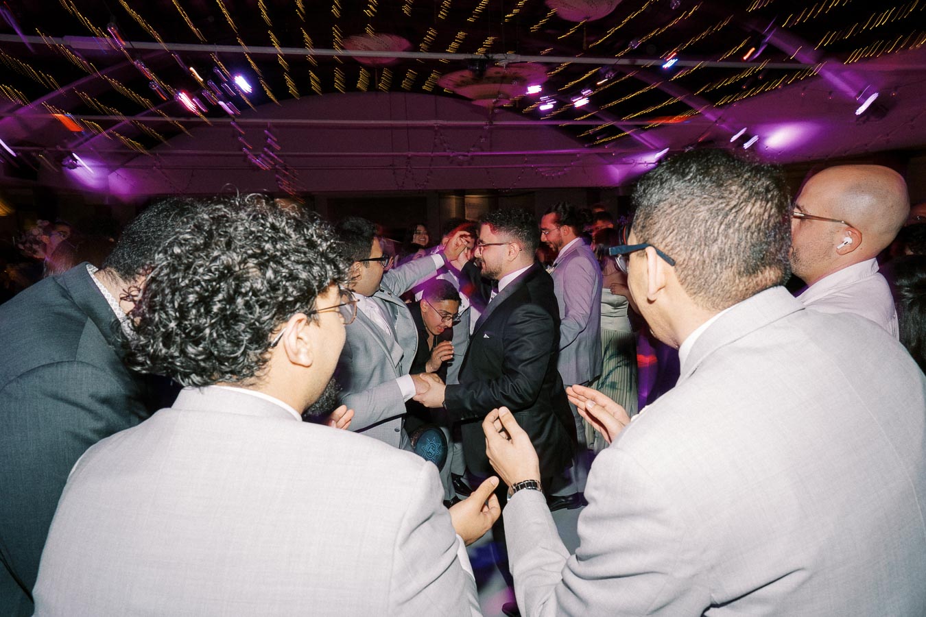 A lively wedding reception with guests in suits and ties dancing under colorful lights, creating a festive atmosphere.