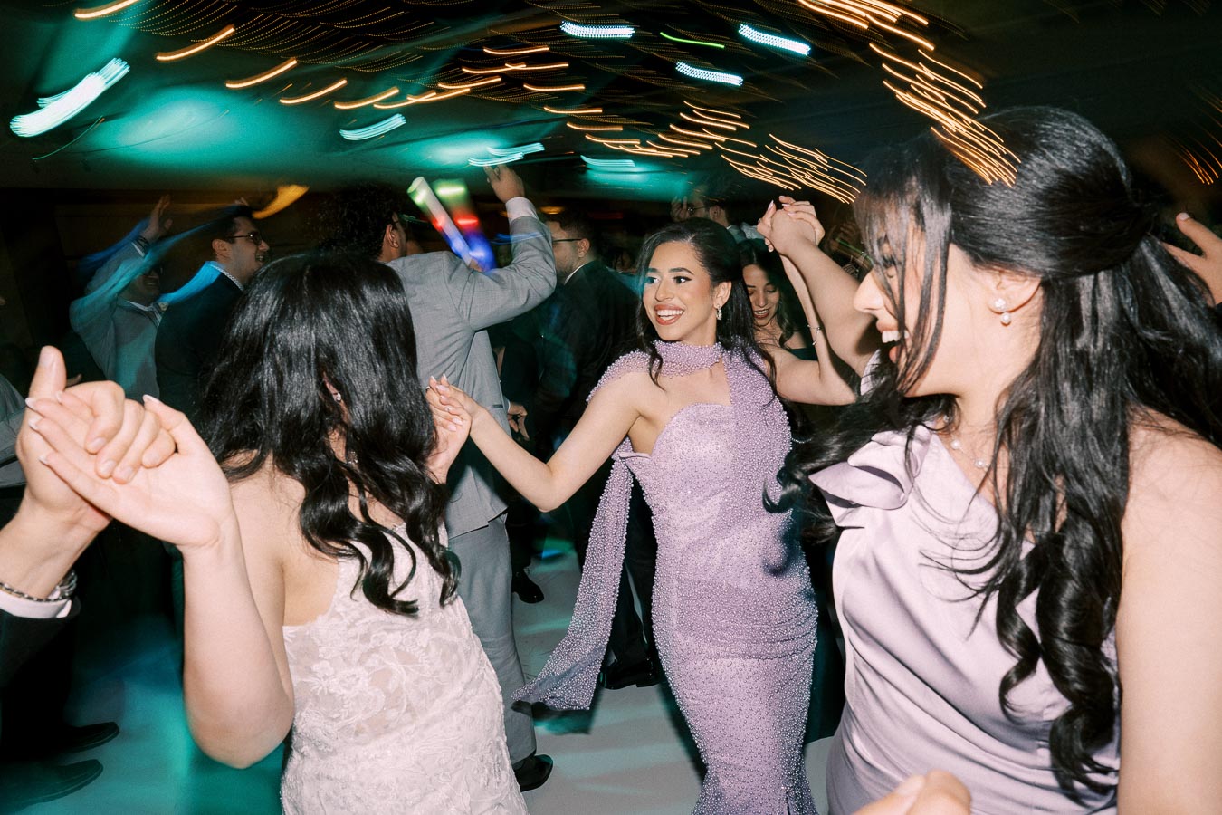 Group of people joyfully dancing at a festive event, with vibrant lights and elegant attire creating a lively atmosphere.