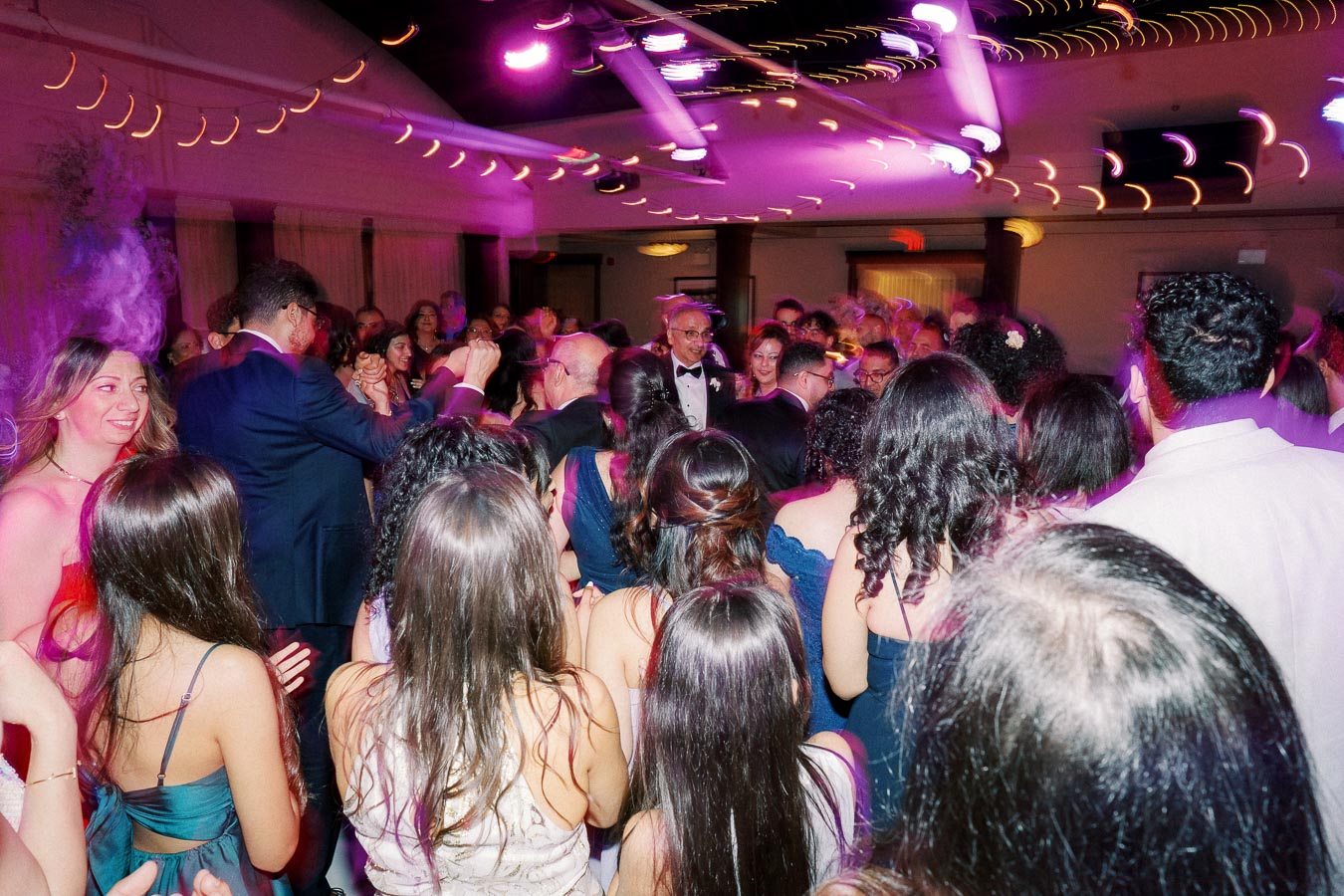A lively party scene with a large group of people dancing and enjoying music under vibrant purple lighting, captured at an indoor event venue.