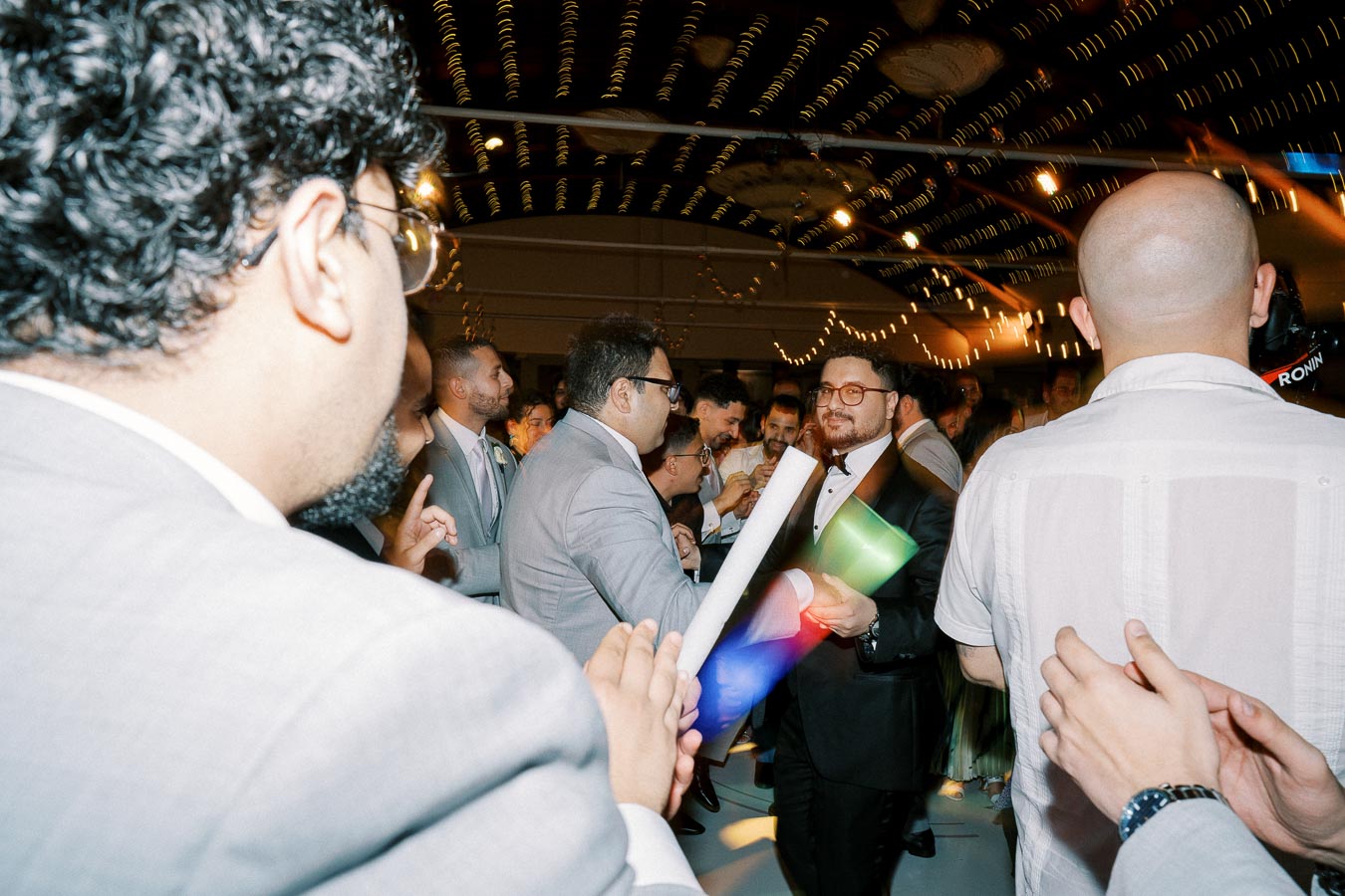 A lively celebration with a crowd of guests in formal attire, gathered in a decorated venue with string lights overhead, as two people greet each other holding scrolls and colorful cylinders.