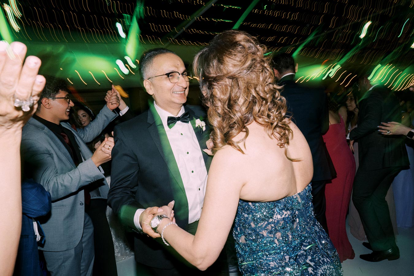 A couple dancing at a lively wedding reception, surrounded by guests in formal attire under vibrant green and yellow lighting effects.