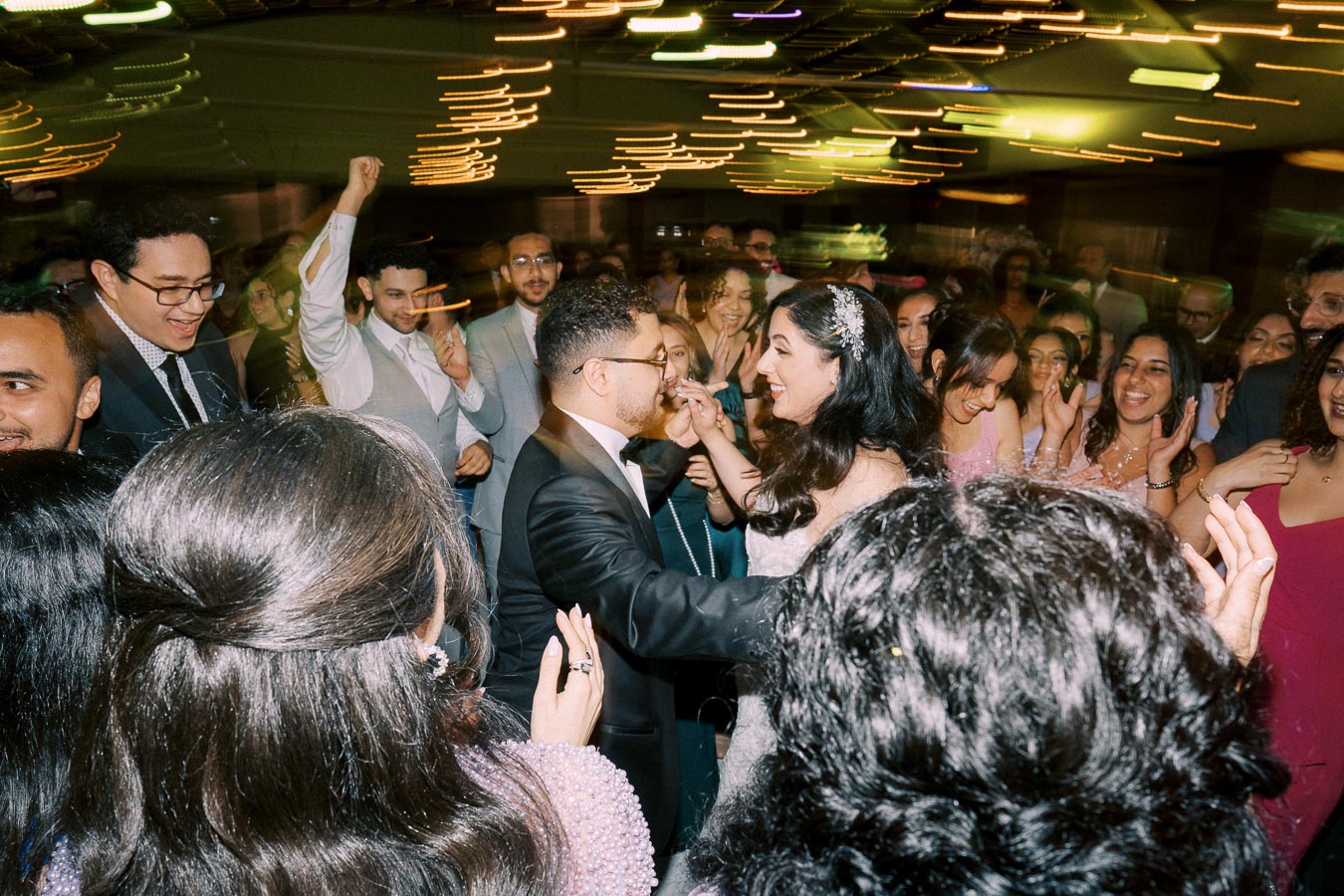 A joyful wedding celebration with a bride and groom dancing together, surrounded by guests in festive attire, capturing a vibrant and lively atmosphere with motion blur and colorful lighting.