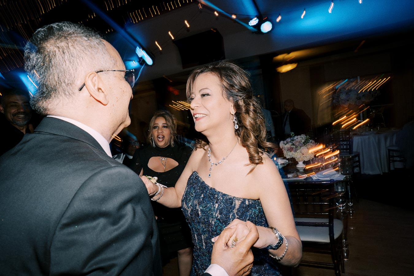 A smiling couple dancing at an elegant event, with guests and decorative lighting in the background.