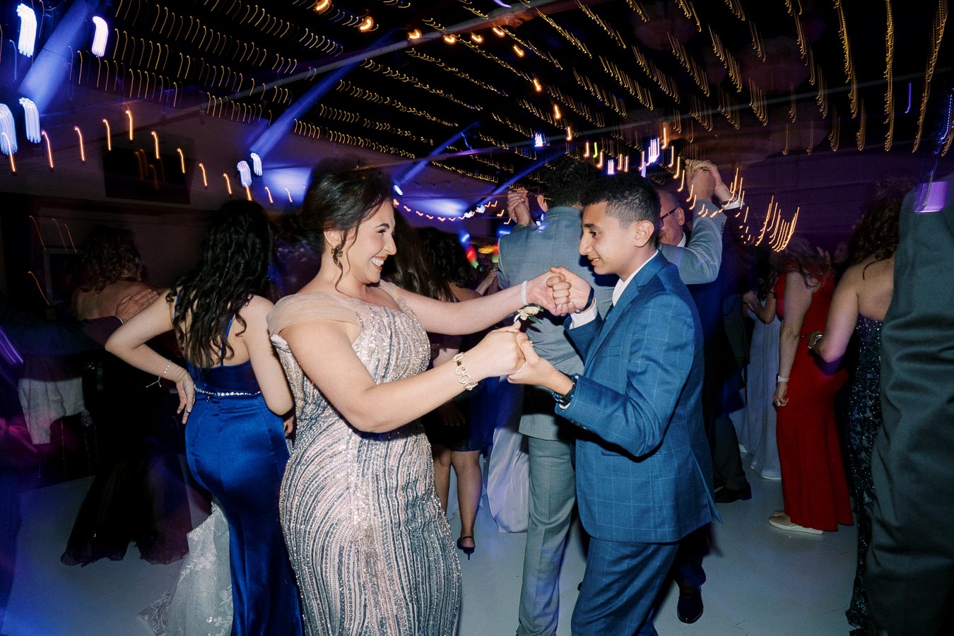 A young couple in formal attire enjoying a lively dance at a prom with vibrant lights and a festive atmosphere.