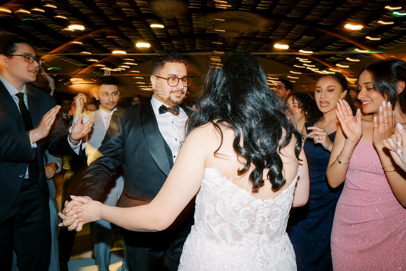 A joyful wedding celebration with a bride in a white lace gown and groom in a black tuxedo dancing, surrounded by smiling guests applauding under warm, ambient lighting.