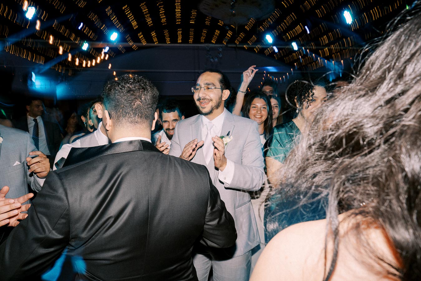 A joyful group of people dancing and celebrating at a lively wedding reception, dressed in formal suits and dresses, under a ceiling adorned with bright, festive lights, creating a vibrant party atmosphere.