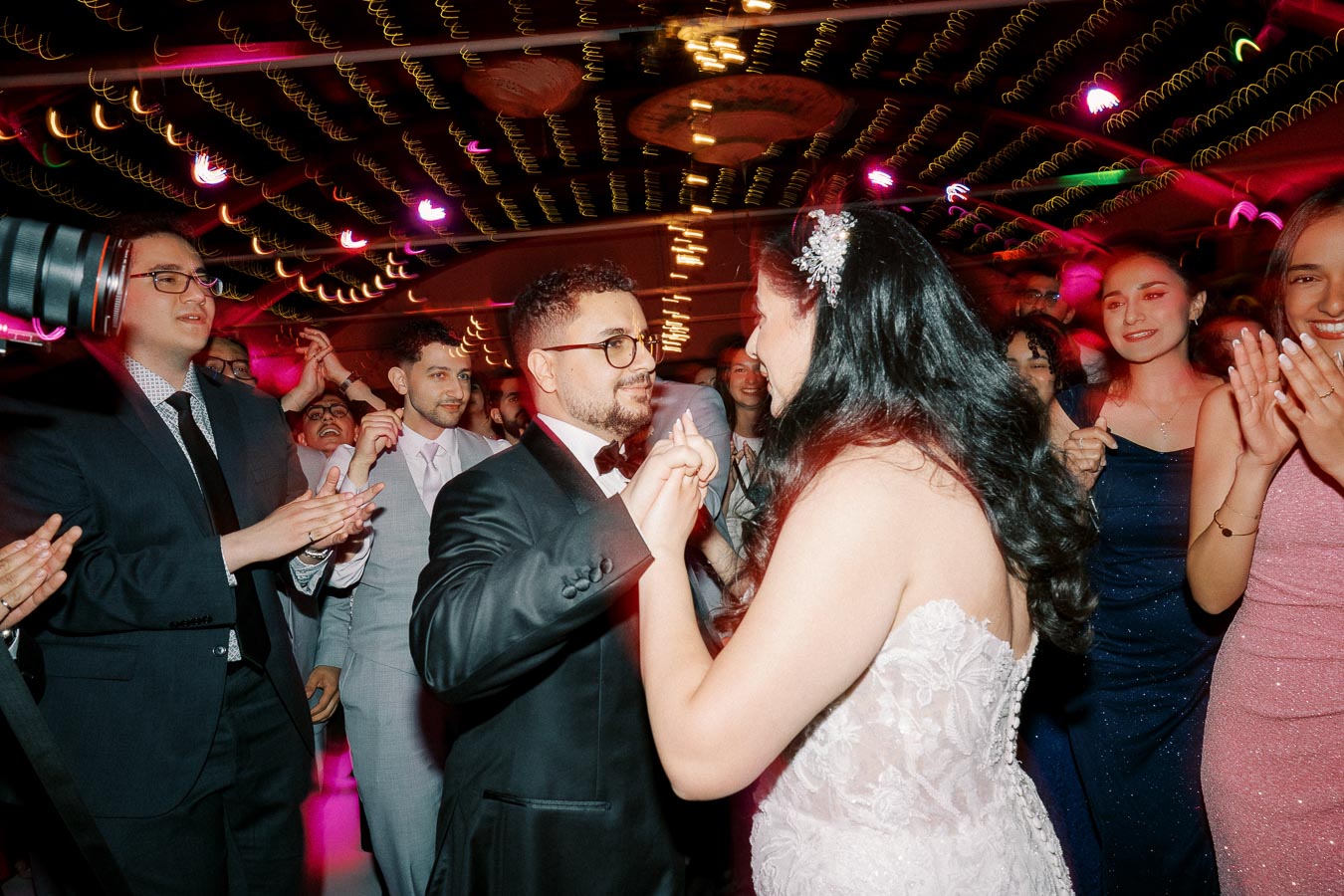 A newlywed couple dancing surrounded by friends in formal attire at a lively wedding reception with colorful, festive lighting.