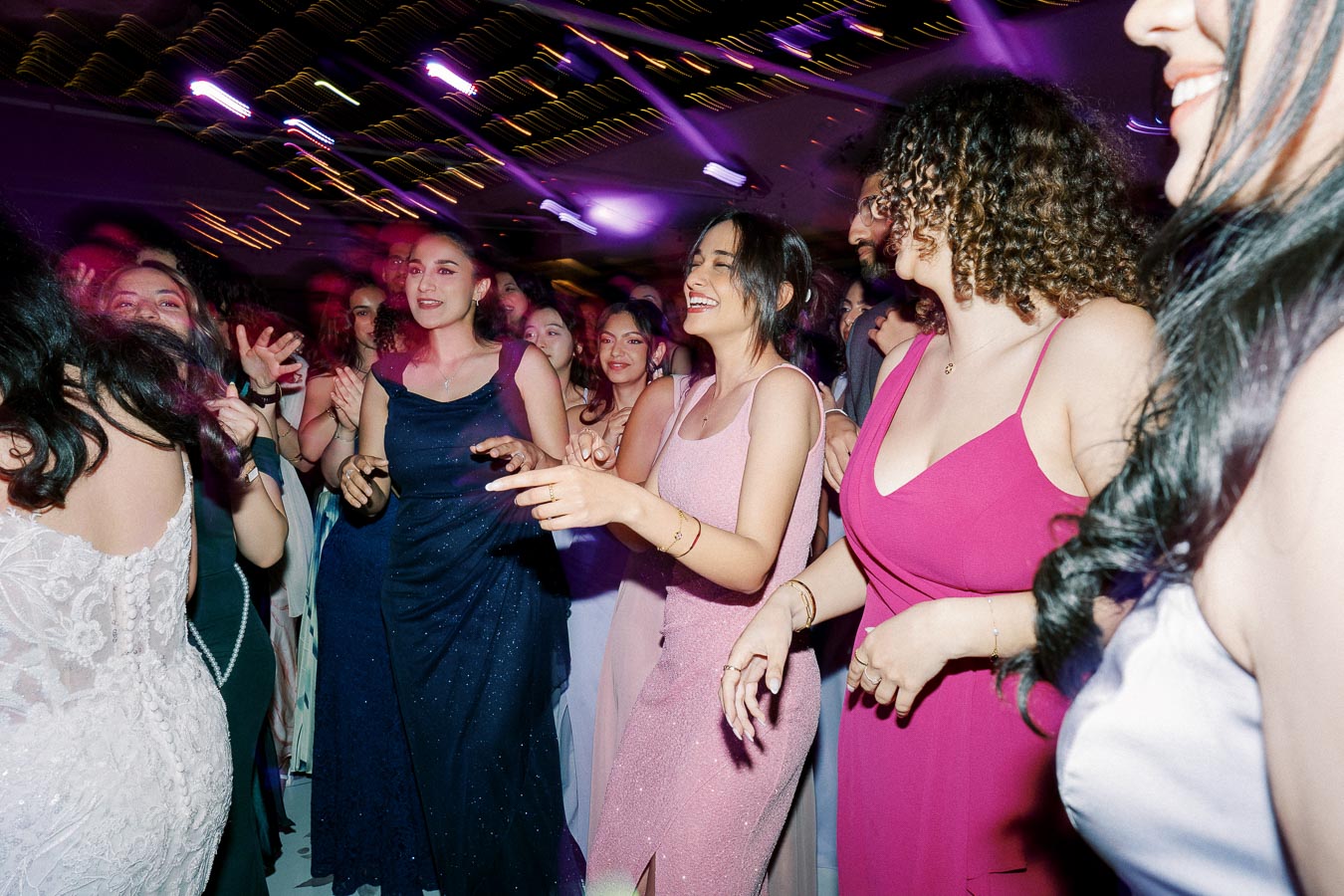 A group of people joyfully dancing at a lively party, wearing elegant evening dresses under vibrant purple lights.