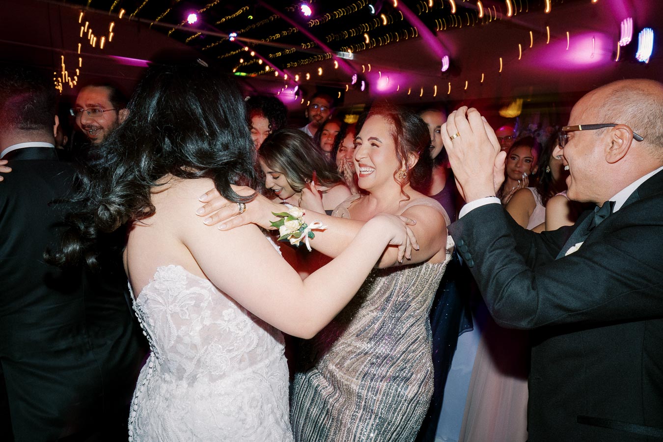 A group of people celebrating and dancing at a wedding reception under vibrant colorful lights. The scene captures joyful expressions, elegant attire, and lively atmosphere.
