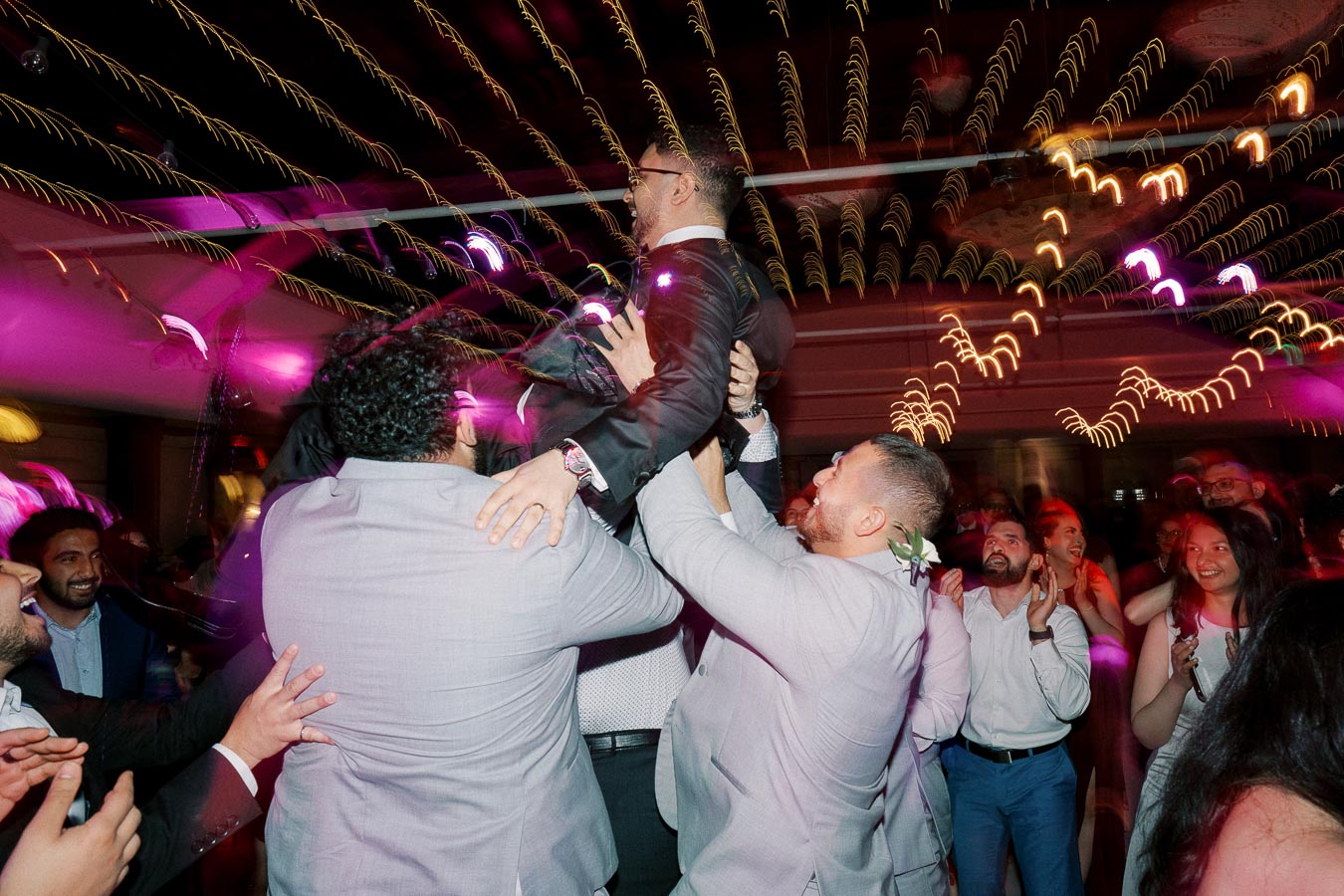 Wedding celebration with guests lifting a man in a suit, under colorful lights and festive atmosphere.