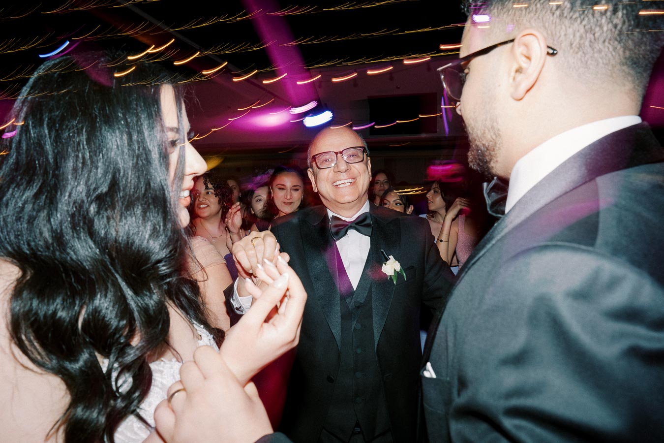 A joyful wedding celebration with guests dancing under colorful lights, featuring a bride and groom holding hands, and a smiling man in a tuxedo.