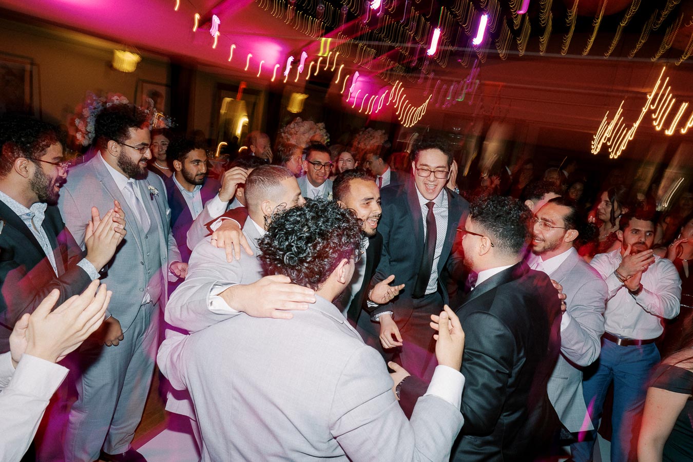 A group of people in suits and formal attire dancing and celebrating under vibrant pink and purple lights at a lively event reception.