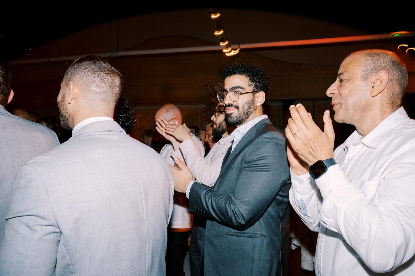 A group of people dressed in formal attire, including suits and shirts, clapping enthusiastically at an indoor event under warm lighting.