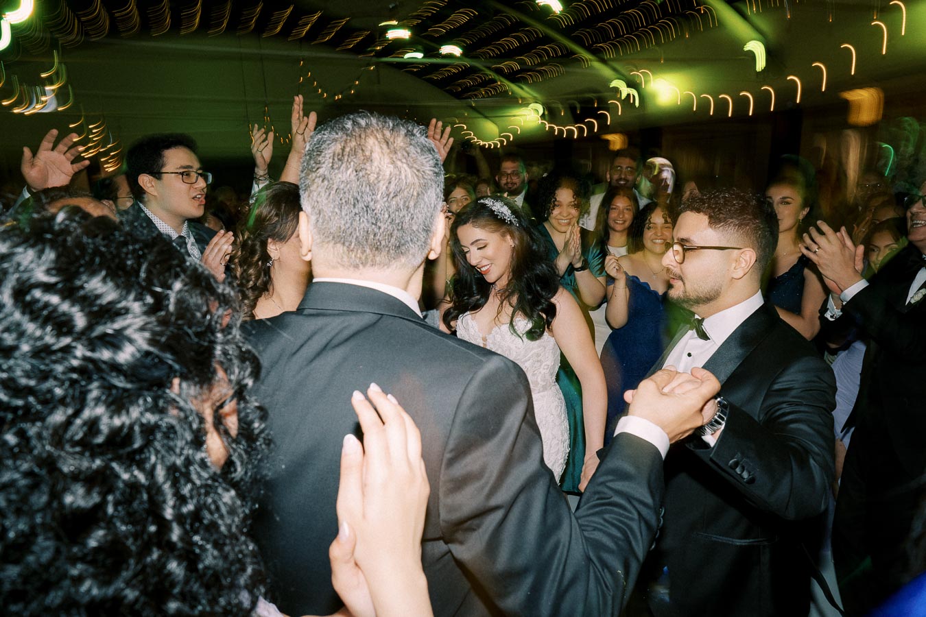 A lively wedding reception with guests dancing, including a smiling bride in a white gown surrounded by elegantly dressed friends and family under festive lights.