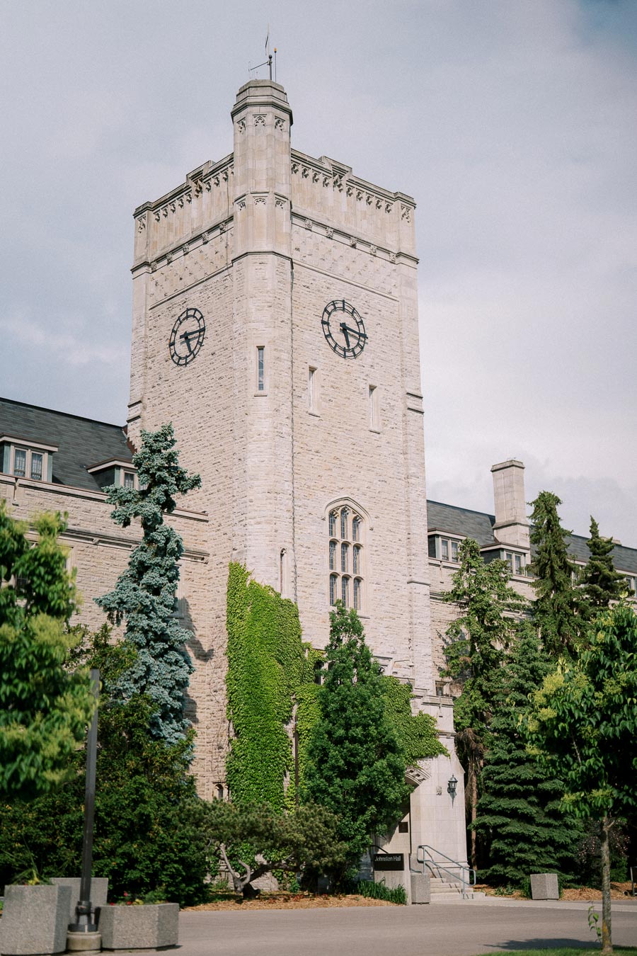 Historic stone building with clock tower surrounded by lush greenery on a sunny day.
