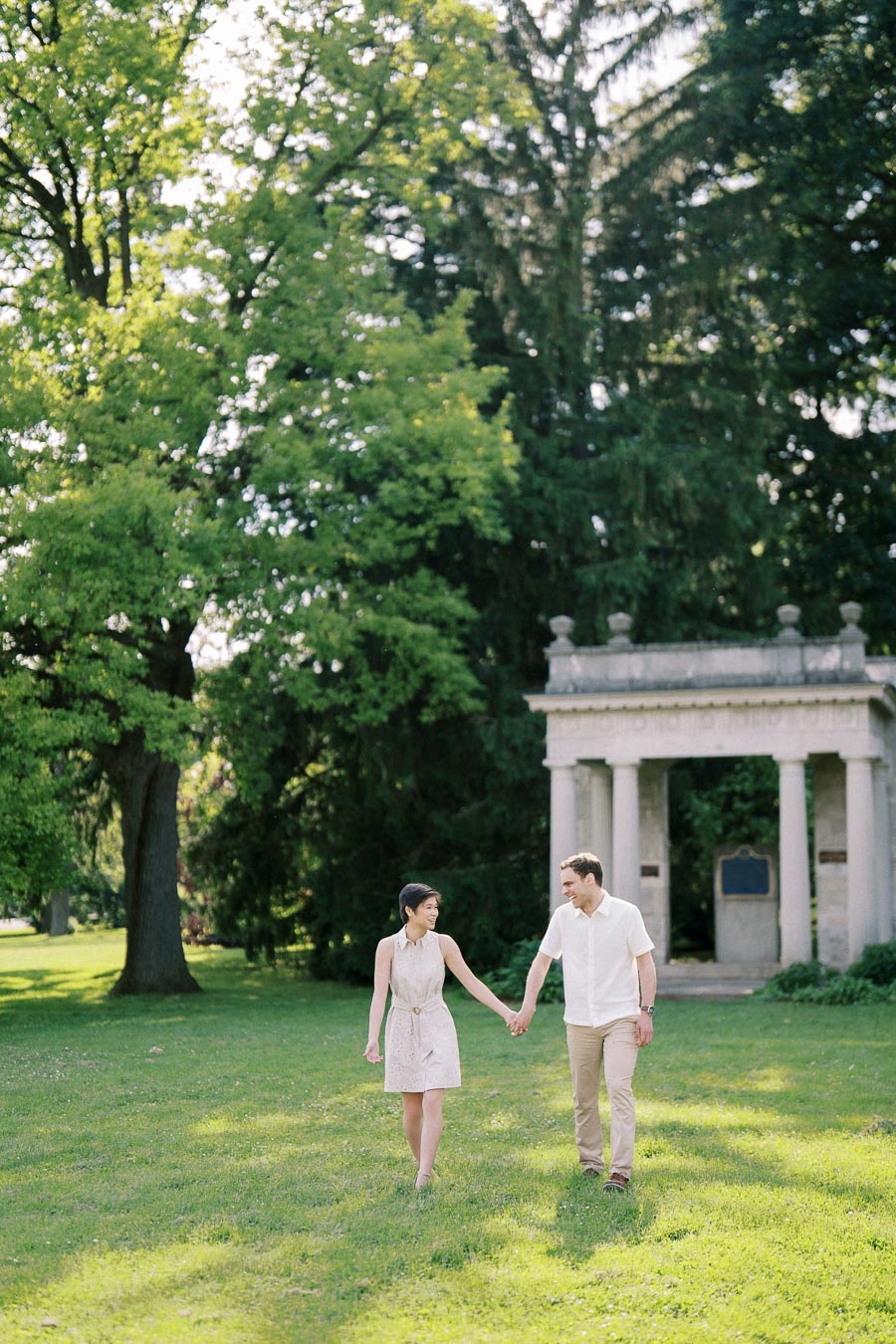 A couple holding hands and walking on a sunny day in a park with green grass and large trees, near a stone structure.