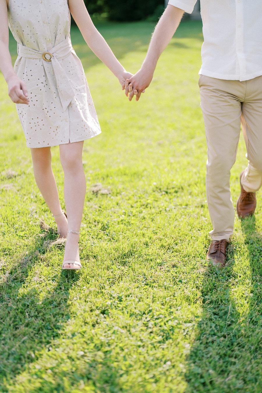 A couple holding hands and walking on a sunlit grassy field, showcasing a light, summery outfit of a beige dress and trousers, embodying a romantic outdoor moment.