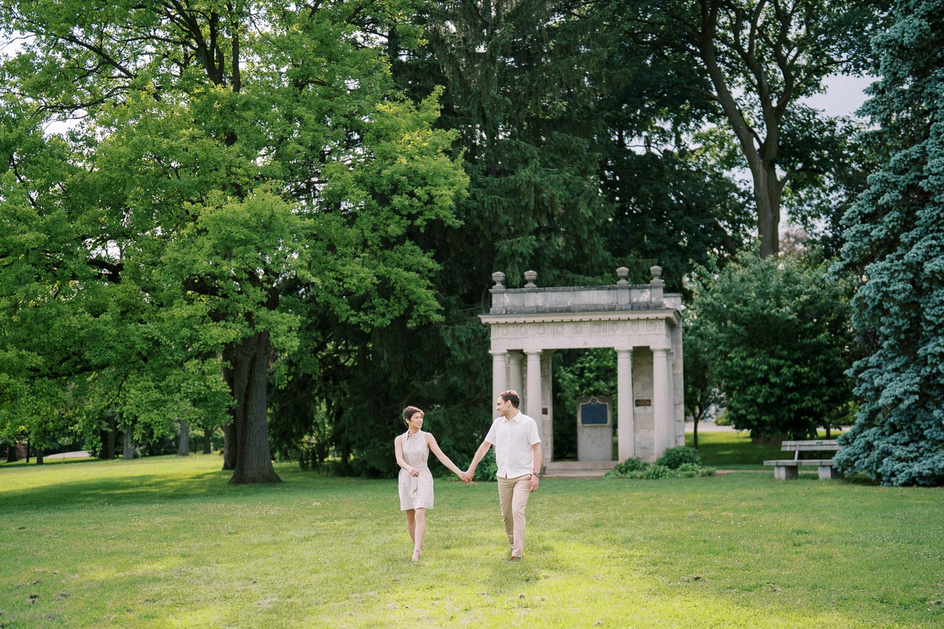 A couple holding hands and walking through a lush green park with tall trees and a classic stone archway in the background, capturing a romantic outdoor moment.