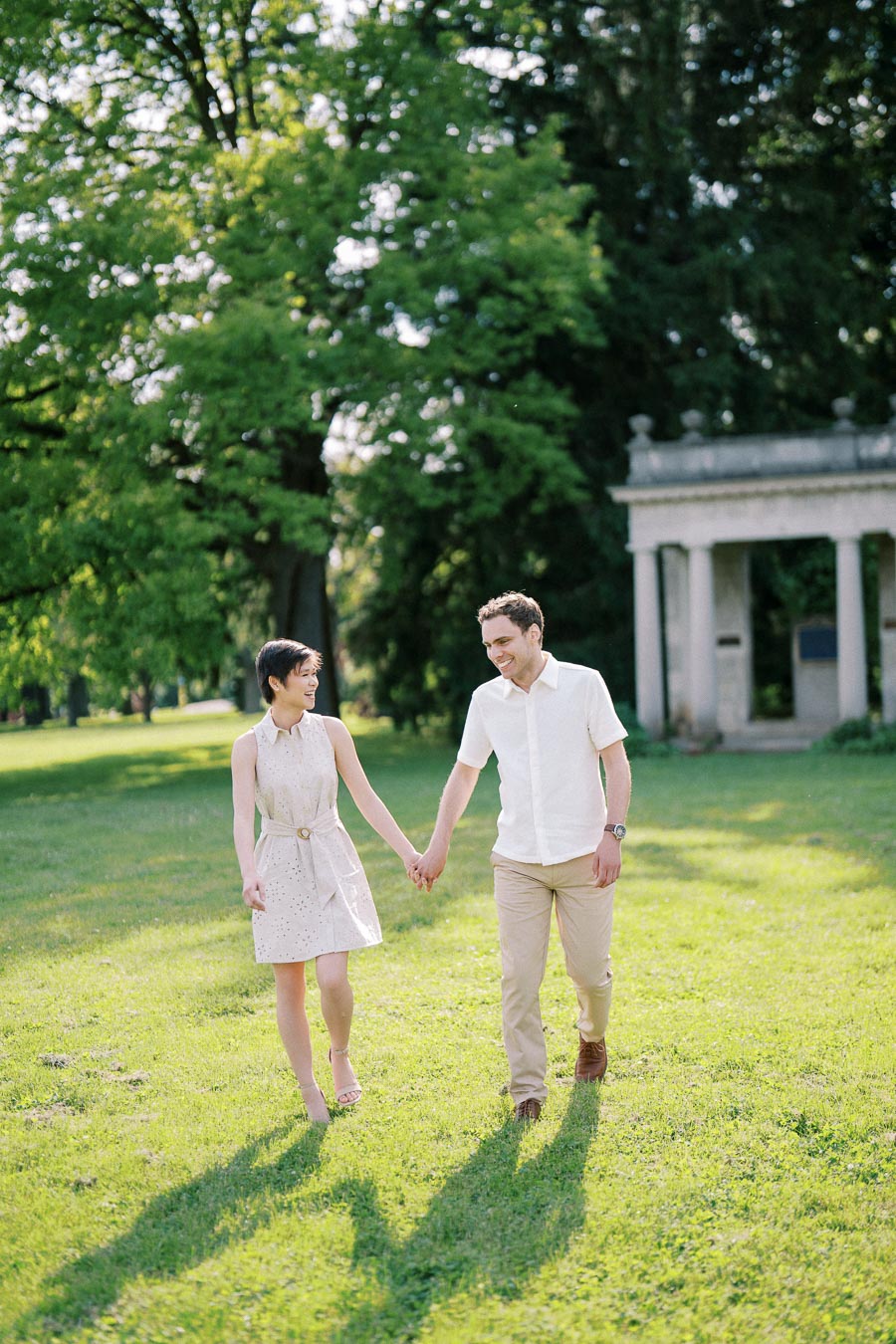 Couple walking hand in hand on a sunny day in a scenic park with lush greenery and a classical stone structure in the background.