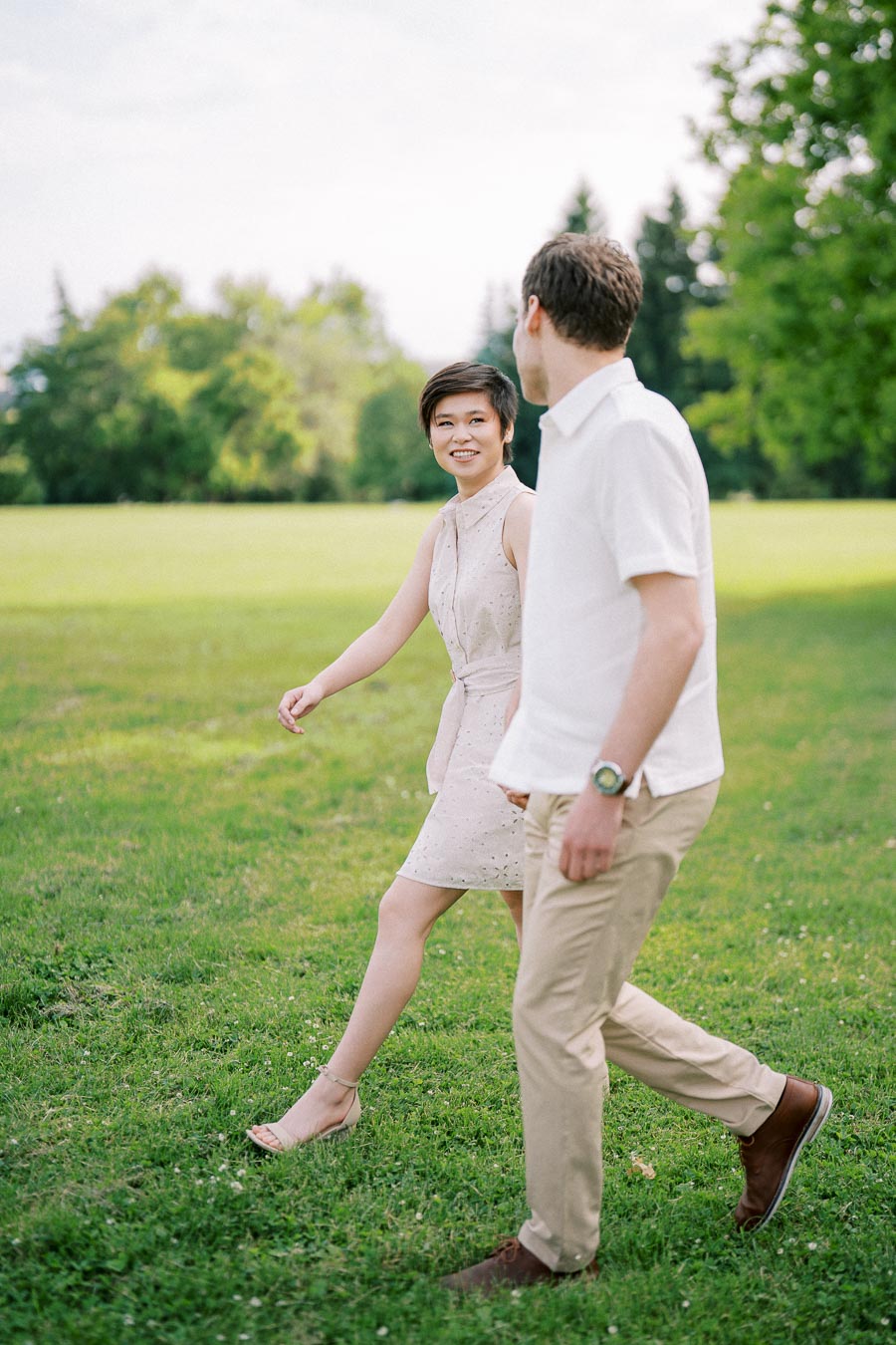 A couple walking and smiling in a lush green park on a sunny day, with trees in the background.