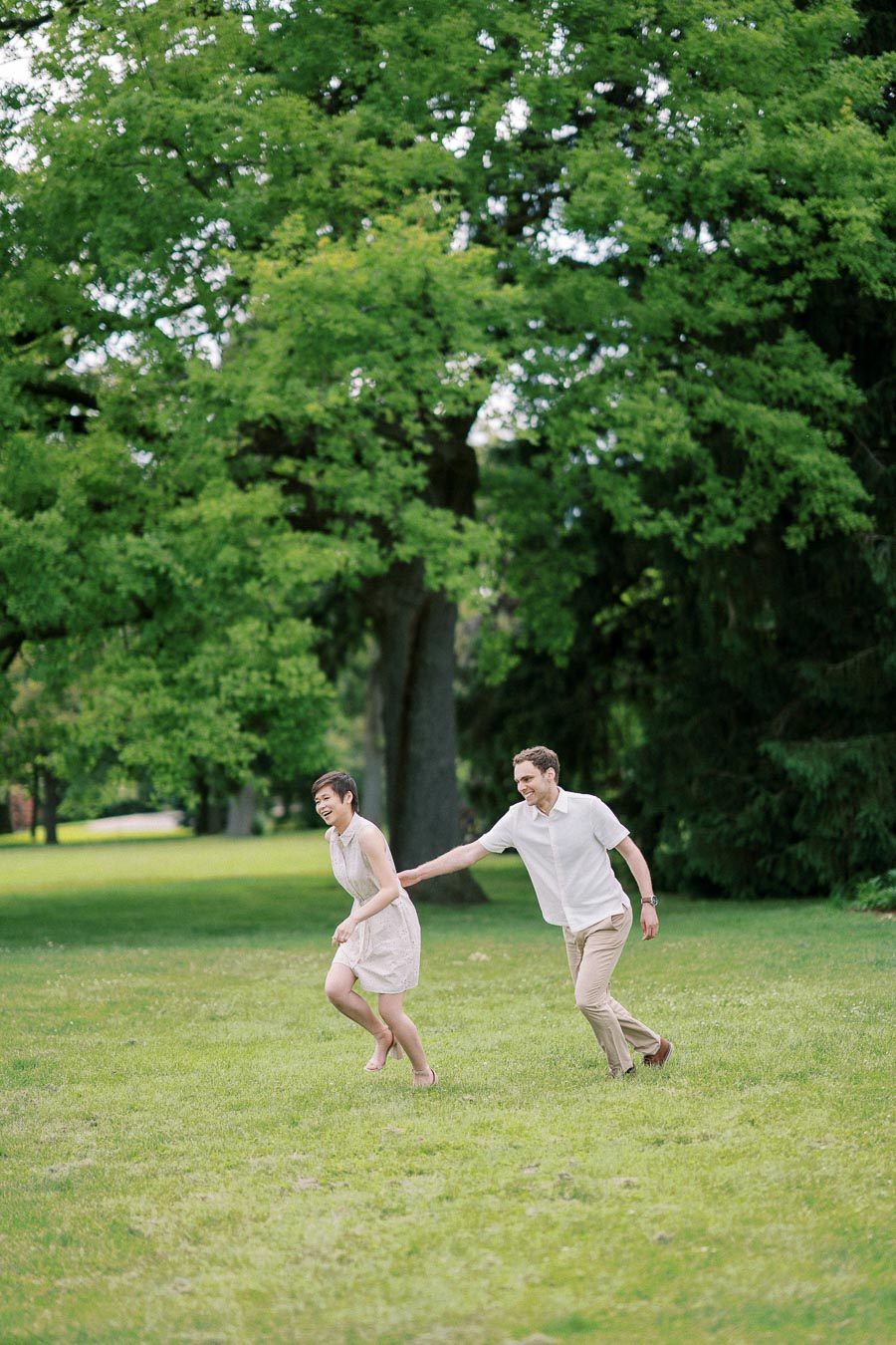 A joyful couple playing and running on a lush green lawn, surrounded by tall trees in a park setting, under a clear sky.