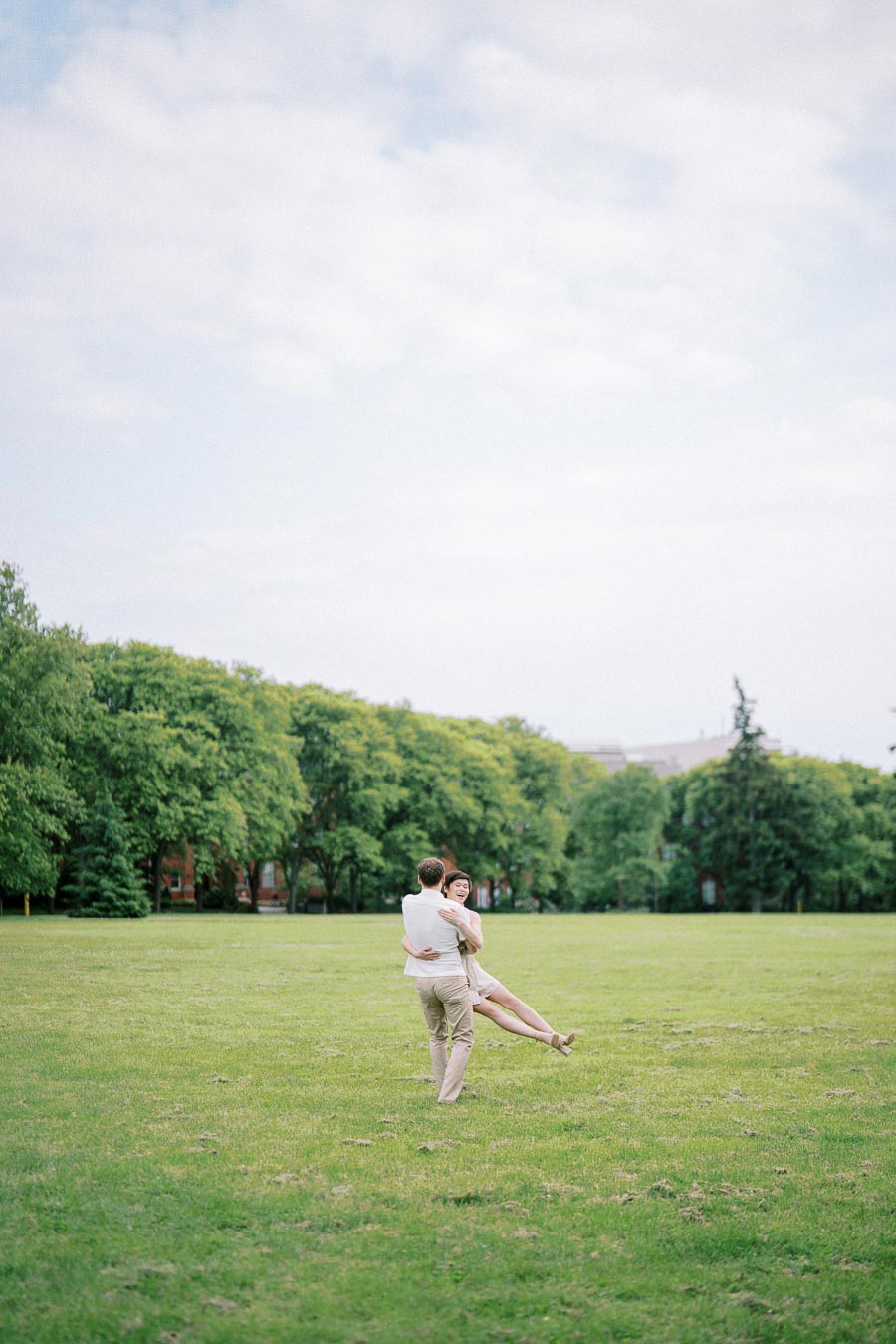 A couple embracing joyfully in a lush green park, surrounded by tall trees under a partly cloudy sky.